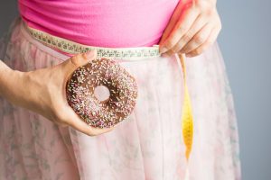 Woman holding donut in hand and check out his body fat with measuring tape.